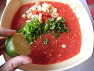 Tomato Watermelon Gazpacho mixture in a large mixing bowl, topped with chopped cucumber, parsley, tomato and jalapeno, with a half of lime being squeezed on top 