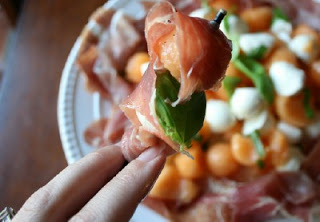 A hand holding a food pick with melon, basil and prosciutto 