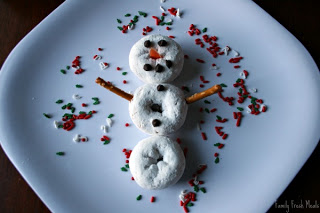 Snowman made of powdered mini donuts, pretzels and mini chocolate chips, on a white plate, surrounded by holiday food sprinkles