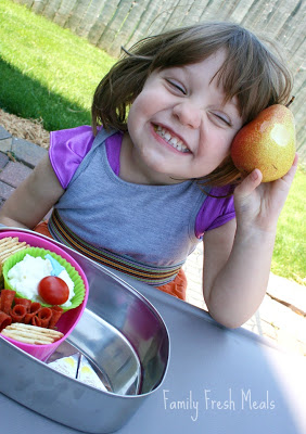 child sitting at a table with lunchbox and holding a pear