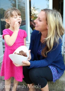 woman holding a bowl of Butterfinger Muddy Buddies in a white bowl with a child eating some chex mix.