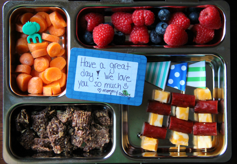 top down image of planetbox lunchbox packed with Turkey sausage and cheese skewers, fresh blueberries and raspberries, carrots, and Butterfinger Muddy Buddies.