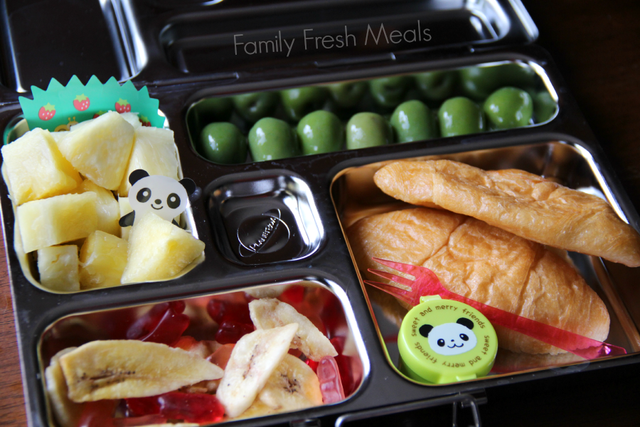 School lunch packed with 2 mini croissants, a small container of jelly, olives, pineapple, banana chips and some fruit snacks.