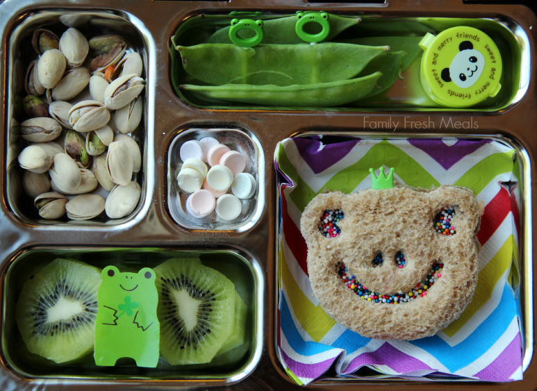 pistachios, snap peas with a container of dip, a frog shaped sandwich, kiwi and a couple candies packed in a metal lunchbox