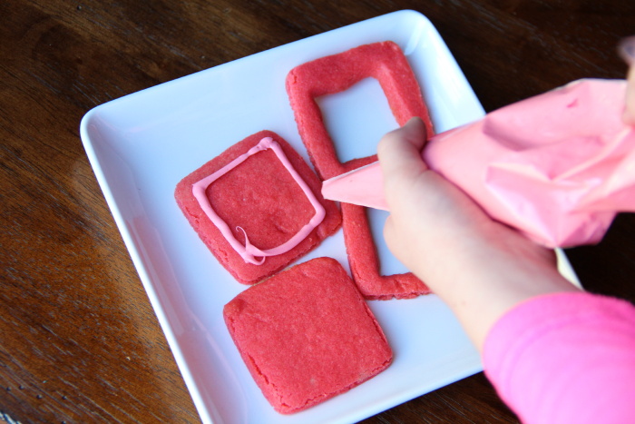 applying pink frosting with pipping bag to red square cookies