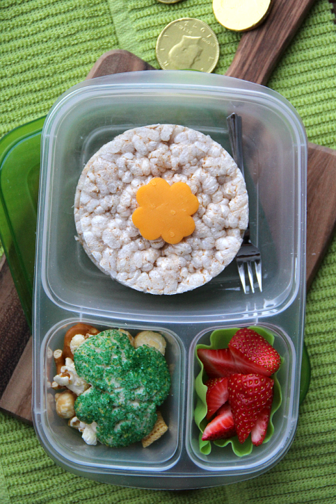 Lunch box packed with a rice cakes topped with a cheese, fresh strawberries, snack mix and a clover sugar cookie.