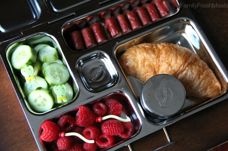 In a metal lunch box - mini croissant, a small container of jelly, turkey sausages, cucumbers, and some fresh raspberries. 