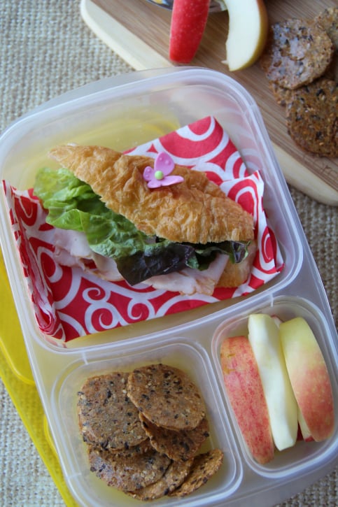 Croissant sandwich, with some apple slices, and crackers packed in a lunch box