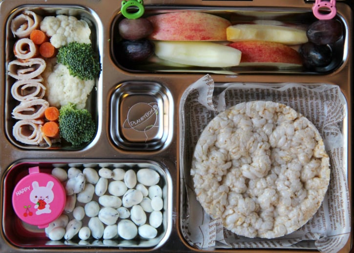 rice cake, apples, grapes, turkey rolls, fresh veggies with a container of dip, and yogurt covered raisins - all packed in a metal lunchbox