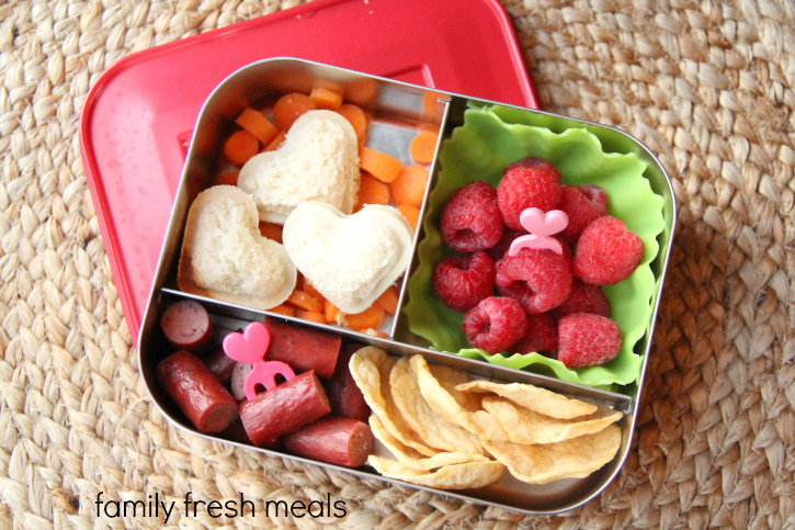 Mini heart sandwiches packed in a small metal lunchbox with chips, carrots, raspberries and mini sausages