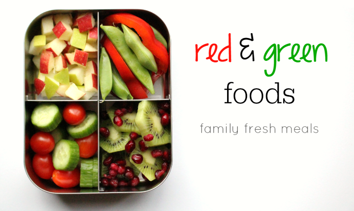top down image of a lunchbox with green and red foods - apples, peppers, edamame, tomatoes, cucumbers, kiwi, pomegranate seedss