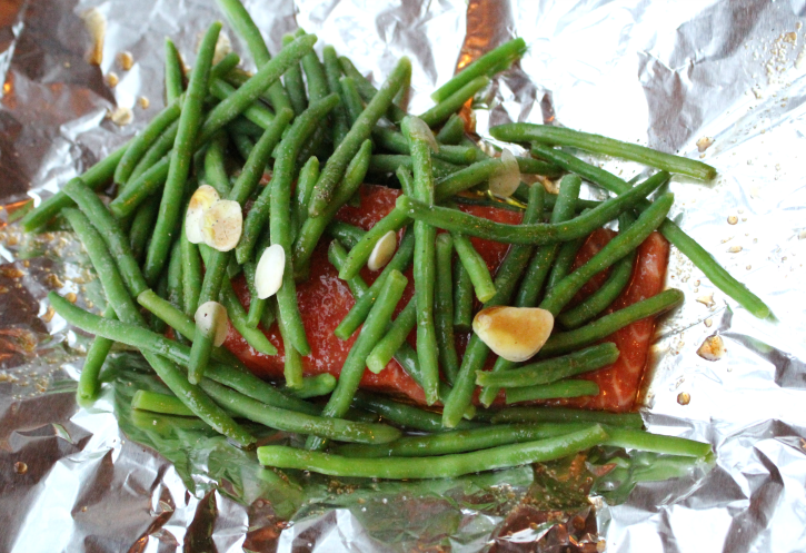 Salmon sitting in the middle of foil, topped with green beans, garlic slices and a brown sauce