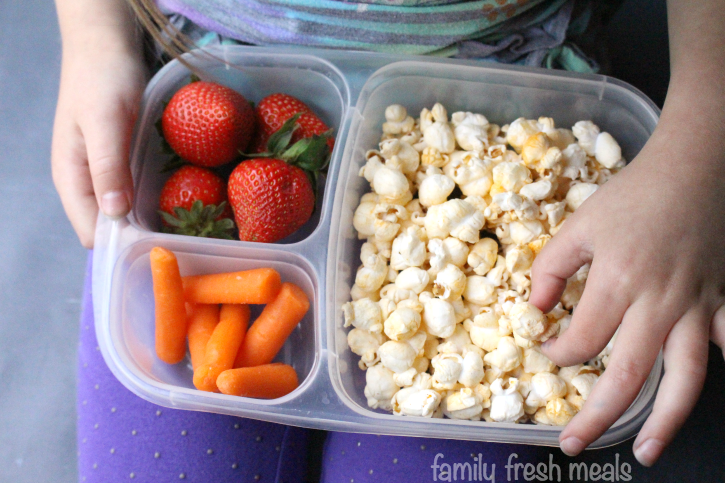 child hold lunchbox on lap, packed with popcorn, strawberries and carrots