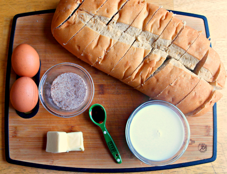 Ingredient for Overnight Snickerdoodle French Toast on a cutting board: loaf of bread, eggs, cinnamon sugar, vanilla, cream and butter