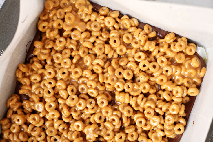 No Bake Chocolate Caramel Cereal Bars - Caramel Cheerios spread on top of chocolate in a white baking dish