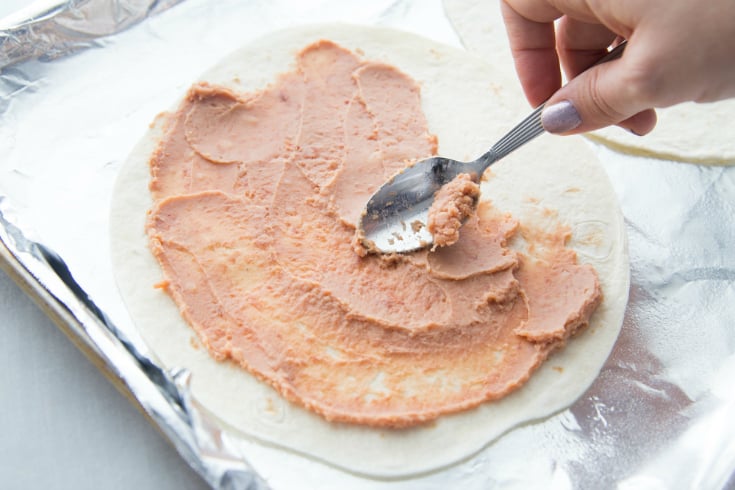 Copycat Taco Bell Mexican Pizza Stacks - Spreading refried bean on tortilla with a spoon