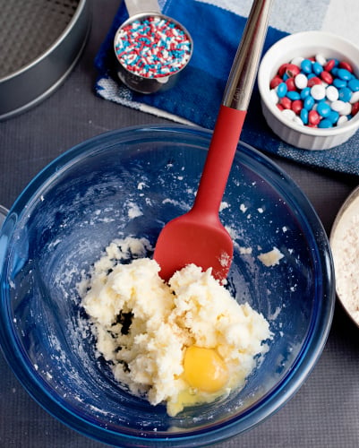 Sugar Cookie Cake - dough and egg in a glass mixing bowl. Red white and blue m&ms and sprinkles sitting next to the bowl