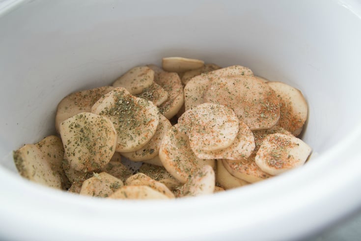 Crockpot Cheesy Cowboy Potatoes - Sliced potatoes in a white crockpot with seasonings