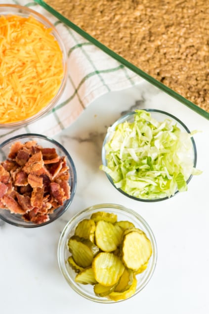 Bacon Cheeseburger Dip - Topping ingredients in small bowls_ bacon, pickles, shredded lettuce and shredded cheese