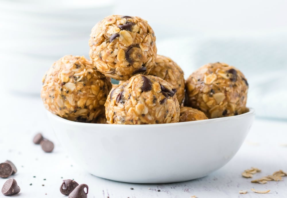 Side photo of Energy bites in a white bowl, surrounded by chocolate chips, and oat pieces