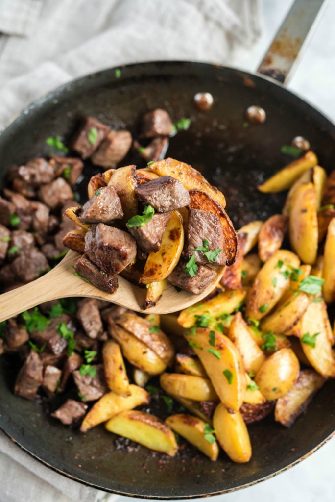 A wooden spoon scooping up steak and potato bites from frying pan