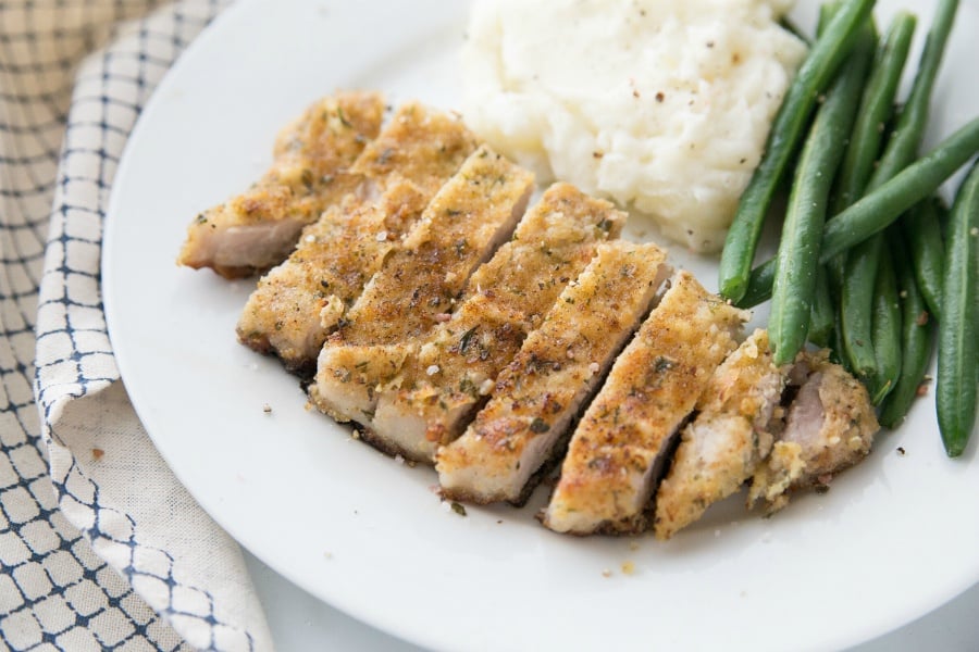 parmesan crusted pork chop, sliced, on a white plate with green beans and mashed potatoes