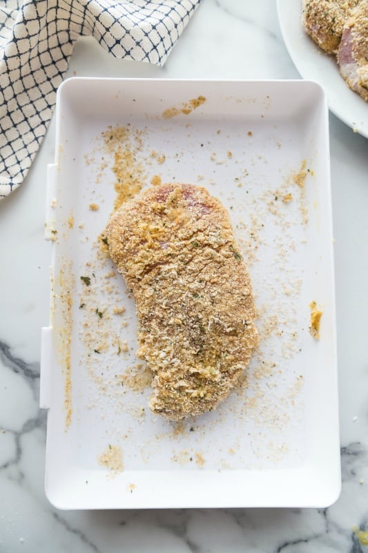 raw pork chop coated with bread crumb parmesan cheese mixture, sitting on a white plate