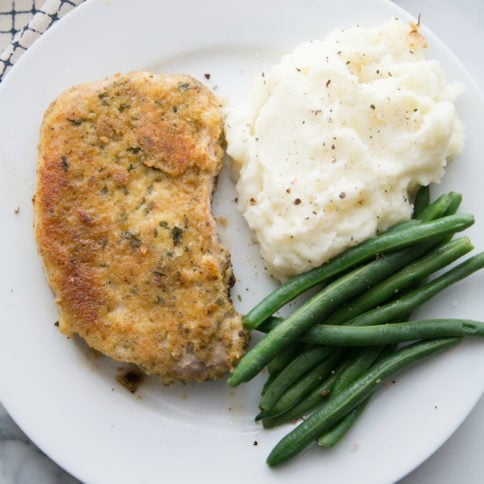 Top down picture of parmesan crusted pork chop on a white plate with green beans and mashed potatoes