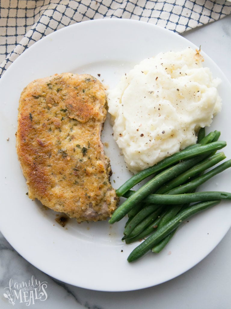 Top down picture of parmesan crusted pork chop on a white plate with green beans and mashed potatoes