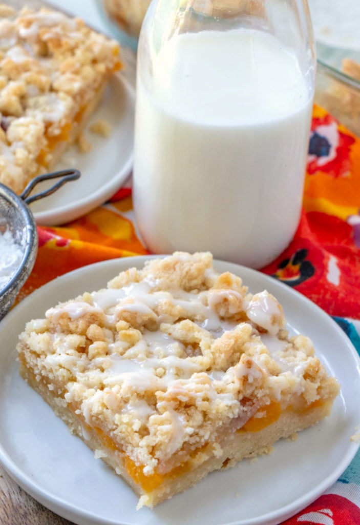 Peach Crumble Bars - piece of peach cumble bar on a white plate with jug of milk in background
