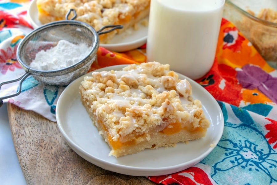 piece of peach crumble bar on a white plate with jug of milk in background and sugar sifter sitting left of the plate