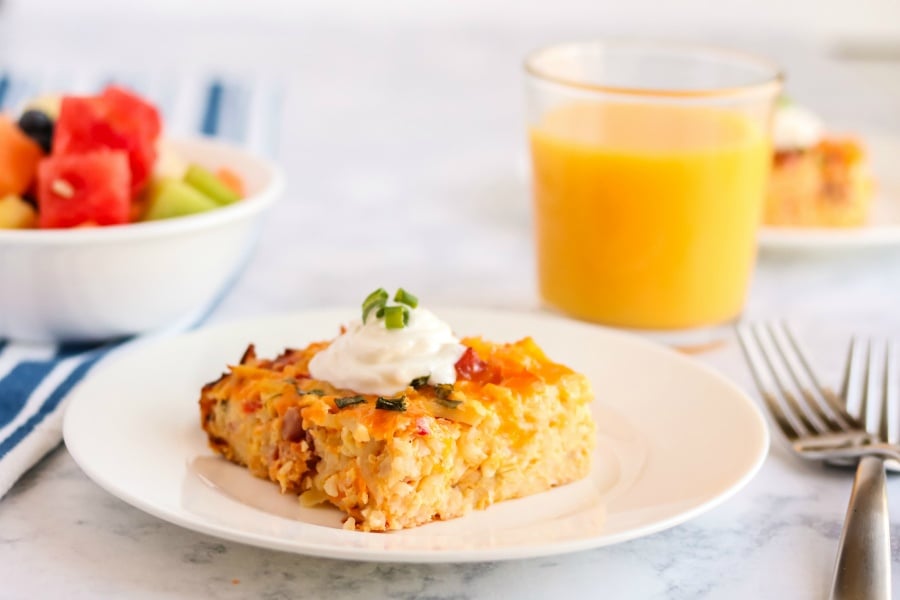 Pimento Cheese Breakfast Casserole - piece of breakfast casserole on a white plate, with a bowl of fruit and a glass of orange juice in the background