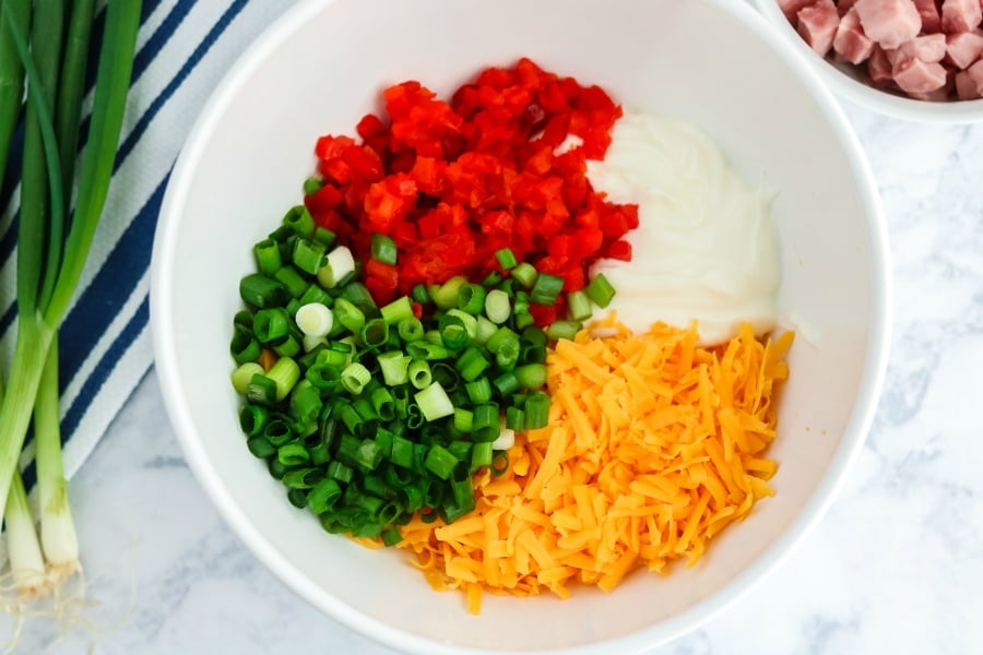 Shredded cheese, chopped green onion, chopped pimentos and sour cream in a white mixing bowl. Small bowl of diced ham in the background