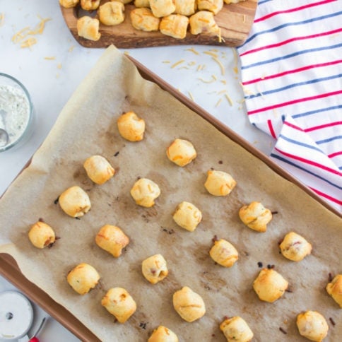 top down image of baked hotdog pieces wrapped in cheesy dough on a parchment paper lined baking sheet and also a pile of hotdog nuggets on a platter