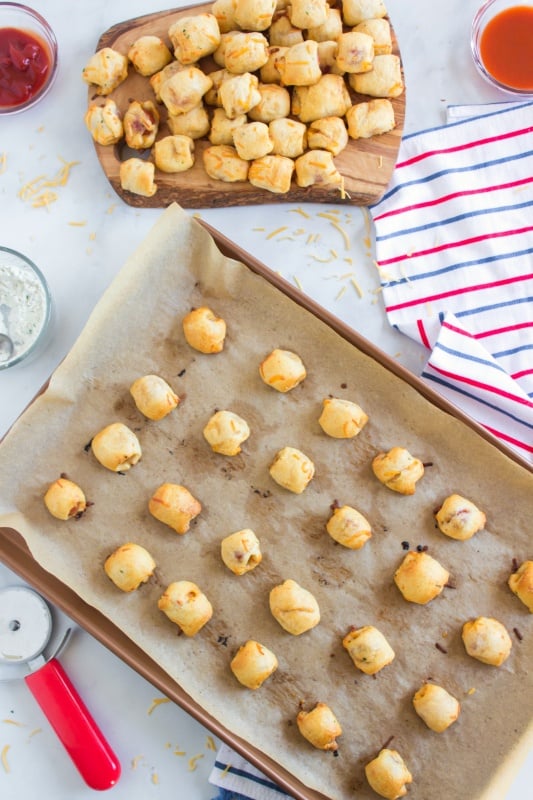 top down image of baked hotdog pieces wrapped in cheesy dough on a parchment paper lined baking sheet and also a pile of hotdog nuggets on a platter