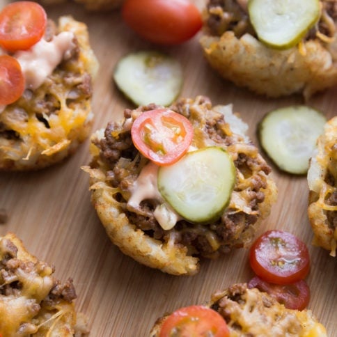 Mini Cheeseburger Tater Tots Cups Topped with pickle and tomato slices, sitting on a cutting board