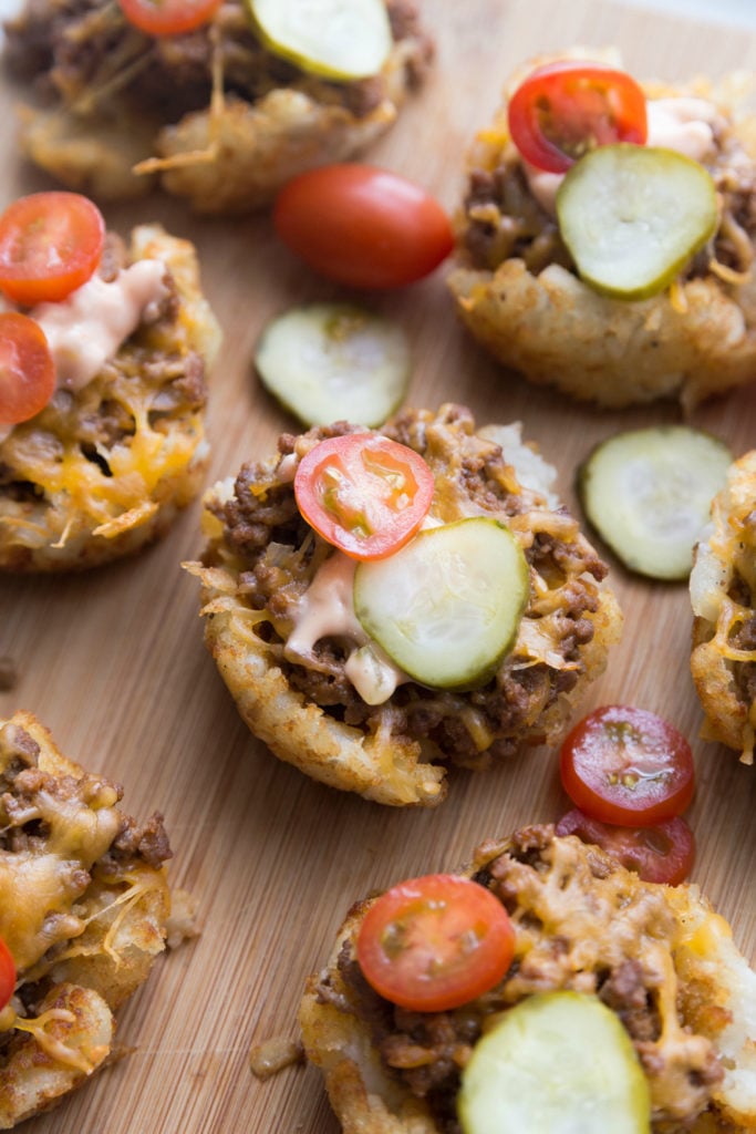 Mini Cheeseburger Tater Tots Cups Topped with pickle and tomato slices, sitting on a cutting board