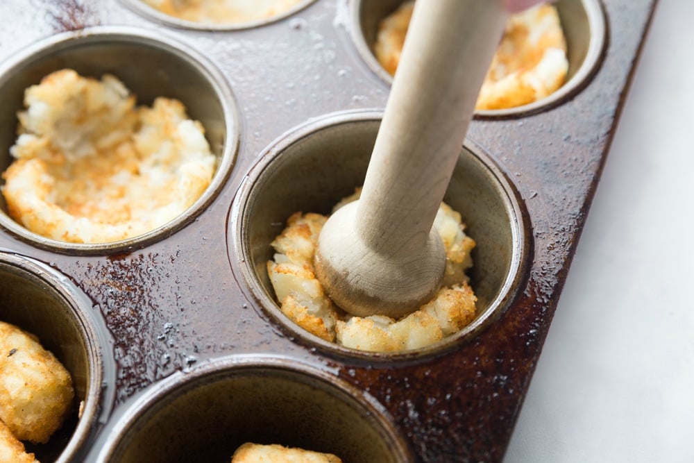 Smashing tater tots down into muffin tin with wooden muller