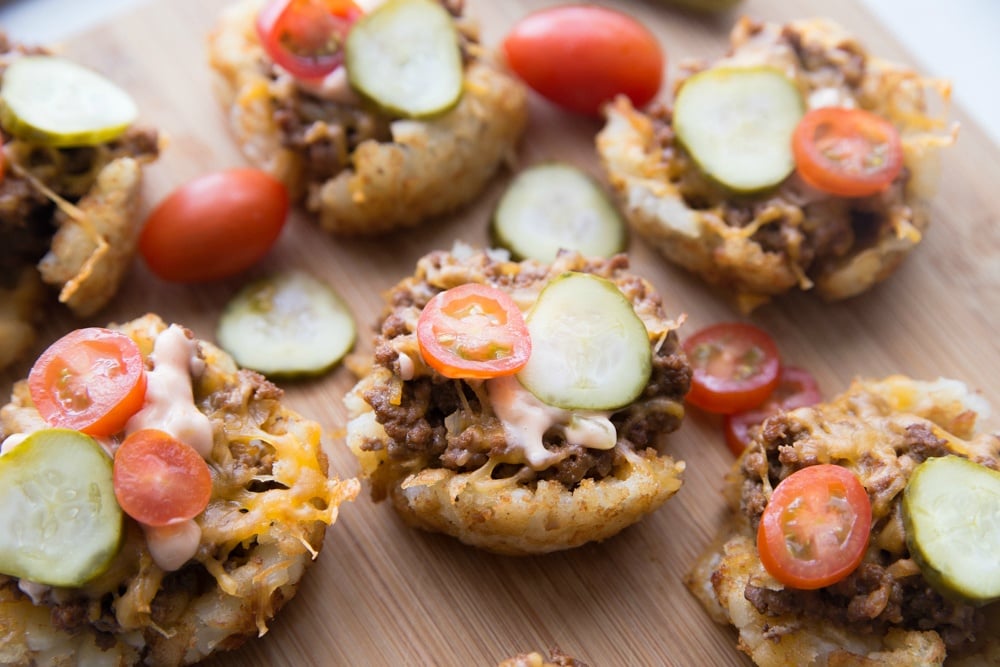Mini Cheeseburger Tater Tots Cups topped with pickle and tomato slices, sitting on a cutting board