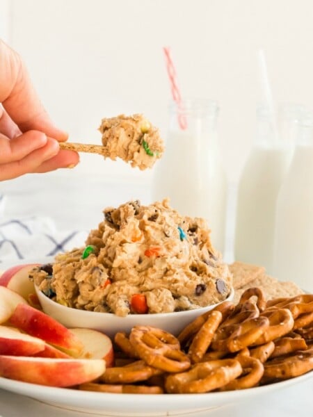 Monster Cookie Dough Dip served in a white bowl, surrounded by pretzels, sliced apples and graham crackers. Glasses of milk in the background