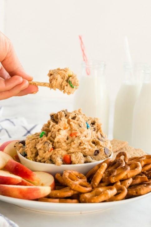 Monster Cookie Dough Dip served in a white bowl, surrounded by pretzels, sliced apples and graham crackers. Glasses of milk in the background