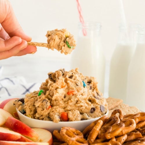 Monster Cookie Dough Dip served in a white bowl, surrounded by pretzels, sliced apples and graham crackers. Glasses of milk in the background