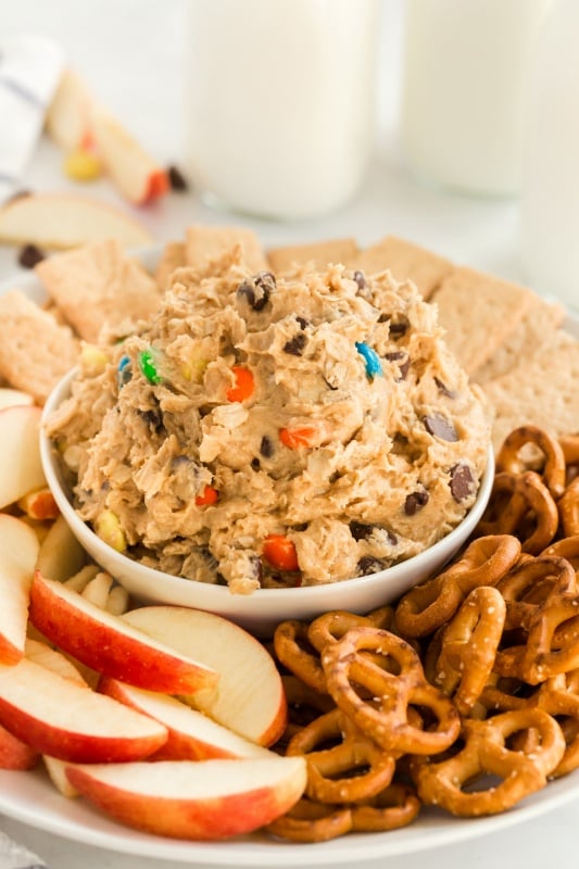 Monster Cookie Dough Dip served in a white bowl, surrounded by pretzels, sliced apples and graham crackers. With glasses of milk in the background