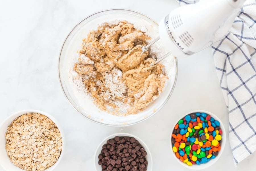 oats, chocolate chips and m and m's in small white bowls. Cookie dough mixture being mixed with a hand mixer