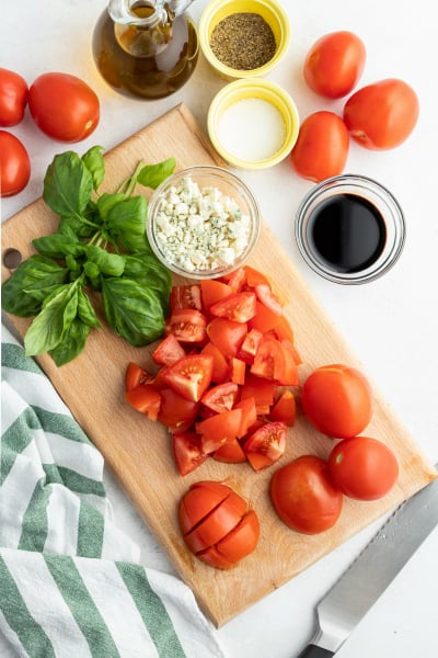 - Ingredients for Balsamic Tomato Salad recipe laid out. Chopped tomatoes on cutting board, along with cheese crumbles, basil, balsamic glaze, salt, pepper and olive oil