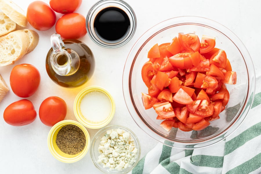 Chopped tomatos in a glass bowl, cheese crumbles, salt and pepper in small bowl, olive oil in a jar and some whole tomatoes off to the side