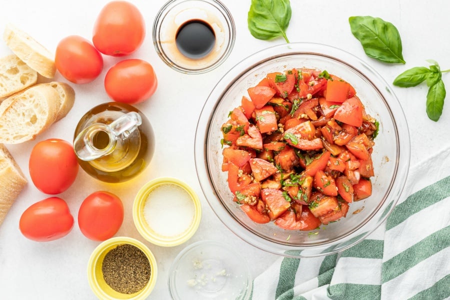Tomato salad all mixed together in a large mixing bowl. Small dishes of salt, pepper, olive oil, whole tomatoes, and frech bread loaf to the left of mixing bowl