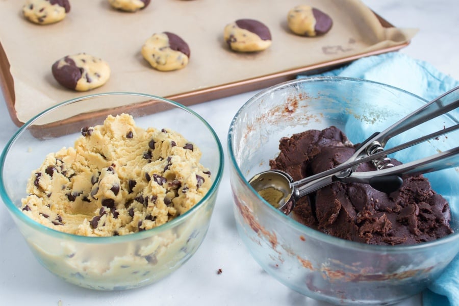 one bowl of chocolate cookie dough and one bowl of brownie batter with a cookie scoop in bowl. Cooked brookies on a baking sheet in background