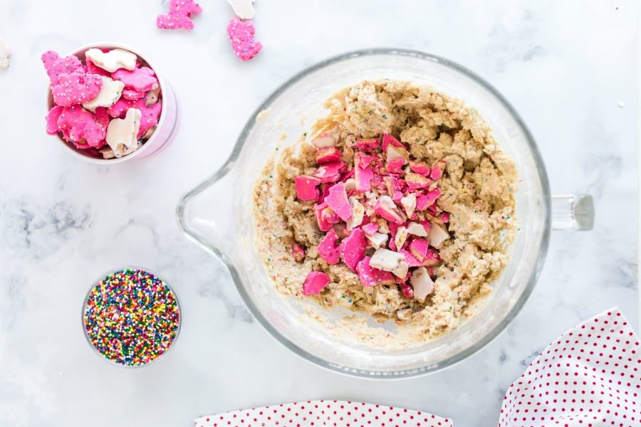 Crushed animal cookies being added to cookie batter in mixing bowl, with a small bowl of cookies, and sprinkles on the side.