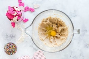 Egg being added to cookie batter in mixing bowl, with a small bowl of cookies, and sprinkles on the side